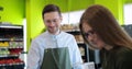 Young cashier in glasses servicing young woman customer in supermarket. Male cashier in green apron at cash desk and Royalty Free Stock Photo