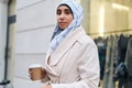 Young casual Arabic woman in hijab walking around street with coffee to go