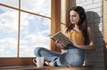 Young casual brunette girl sitting on window sill listening to music and reading a book during quarantine Royalty Free Stock Photo