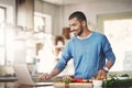 Young casual man in the kitchen on a laptop while preparing a healthy meal at home. Happy smiling male browsing and Royalty Free Stock Photo