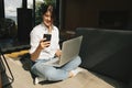 Young casual woman shopping online from home and smiling. Happy stylish girl chatting on phone and using laptop while sitting on Royalty Free Stock Photo