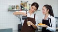 Young caucasian barista pouring milk from the jug into a paper coffee cup. Asian assistant in an apron holding croissant plate Royalty Free Stock Photo