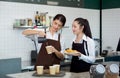 Young caucasian barista pouring milk from the jug into a paper coffee cup. Asian assistant in an apron holding croissant plate Royalty Free Stock Photo