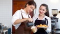 Young caucasian barista pouring milk from the jug into a paper coffee cup. Asian assistant in an apron holding croissant plate Royalty Free Stock Photo