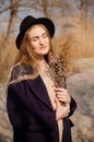 Young caucasian blonde girl in a dress, coat and hat in spring on beach, under rays of sun with willow branches, smiling Royalty Free Stock Photo