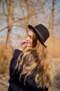Young caucasian blonde girl in a dress, coat and hat in spring on beach, under rays of sun with willow branches, smiling Royalty Free Stock Photo