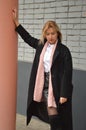 A young Caucasian blonde woman in a black coat, white shirt and boots stands against the background of a wall in the city. Fashion Royalty Free Stock Photo