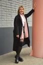 A young beautiful Caucasian blonde girl in a black coat, boots and a pink scarf stands against a brick wall in the city Royalty Free Stock Photo