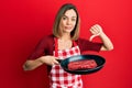 Young caucasian blonde woman wearing cook apron cooking meat on pan with angry face, negative sign showing dislike with thumbs Royalty Free Stock Photo