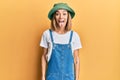 Young caucasian blonde woman wearing denim jumpsuit and hat with 90s style sticking tongue out happy with funny expression Royalty Free Stock Photo