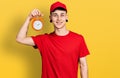 Young caucasian boy with ears dilation wearing delivery uniform and alarm clock looking positive and happy standing and smiling Royalty Free Stock Photo