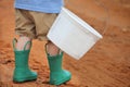 Young caucasian boy walking on a dirt road with a white bucket and wearing green rain boots Royalty Free Stock Photo