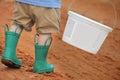 Young caucasian boy walking on a dirt road with a white bucket and wearing green rain boots Royalty Free Stock Photo