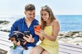 Young caucasian couple using smartphone sitting on the bench with dog at the beach Royalty Free Stock Photo