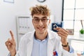 Young caucasian doctor man holding earing aid at the clinic smiling happy pointing with hand and finger to the side Royalty Free Stock Photo