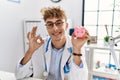 Young caucasian doctor man wearing doctor uniform holding piggy bank at the clinic doing ok sign with fingers, smiling friendly Royalty Free Stock Photo