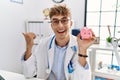 Young caucasian doctor man wearing doctor uniform holding piggy bank at the clinic pointing thumb up to the side smiling happy Royalty Free Stock Photo