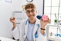 Young caucasian doctor man wearing doctor uniform holding piggy bank at the clinic screaming proud, celebrating victory and Royalty Free Stock Photo