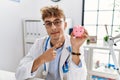 Young caucasian doctor man wearing doctor uniform holding piggy bank at the clinic smiling happy pointing with hand and finger Royalty Free Stock Photo