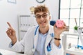 Young caucasian doctor man wearing doctor uniform holding piggy bank at the clinic smiling happy pointing with hand and finger to Royalty Free Stock Photo