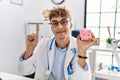 Young caucasian doctor man wearing doctor uniform holding piggy bank at the clinic smiling with an idea or question pointing Royalty Free Stock Photo