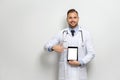 Young caucasian doctor with short hair, mustache and beard dressed in white gown and stethoscope pointing finger at tablet