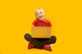 Young caucasian fair-haired student sits on the floor and holds laptop. Boy study at home with internet and computer on yellow