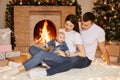 Young Caucasian family posing in festive Christmas room at home while sitting on soft white carpet near fireplace and xmas tree, Royalty Free Stock Photo