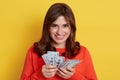 Young Caucasian female with dark hair standing against yellow wall with fan of money in hands, lady wins lottery, having reward,