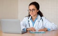 Young caucasian female nutritionist doctor in white coat making notes in clipboard, looking at laptop, sitting at table Royalty Free Stock Photo