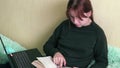 Young caucasian female student in casual clothes is reading a book on the couch at home, holding a laptop on her lap. Student prep