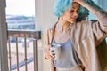 young caucasian female with unusual coloured blue hair on near window with mug