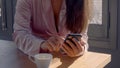 Close up, no face. White girl in a rose robe, with a phone in the hand, drinking at cup of coffee in the sunny morning