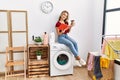 Young caucasian girl drinking coffee and reading book waiting for laundry sitting on whasing machine at home Royalty Free Stock Photo