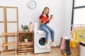 Young caucasian girl drinking coffee and reading book waiting for laundry sitting on whasing machine at home Royalty Free Stock Photo