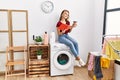 Young caucasian girl drinking coffee and reading book waiting for laundry sitting on whasing machine at home Royalty Free Stock Photo