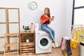 Young caucasian girl drinking coffee and reading book waiting for laundry sitting on whasing machine at home Royalty Free Stock Photo