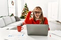 Young caucasian girl sitting on the table working using laptop by christmas tree very happy and smiling looking far away with hand Royalty Free Stock Photo