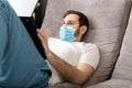 Young caucasian gloomy man working from home office wearing protective mask using laptop and internet. Coronavirus covid 19 Royalty Free Stock Photo