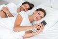 Young caucasian husband sleeping, smiling wife looking at phone, chatting with lover, surfing in internet in bed