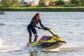 Young Caucasian jet ski woman drives standing on watercraft making splashes at sunset by river bank at countryside. Beautiful