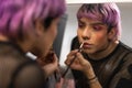 Young caucasian male with purple hair applying lipstick with make up brush looking in the mirror.