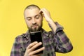 A young caucasian man with a beard and a bald head holding a phone in his hands on a yellow background. Guy holds his head by head Royalty Free Stock Photo