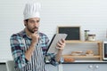 Young Caucasian man with beard wear chef hat and apron holding tablet and put his hands on chin at kitchen