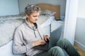 Young caucasian man in casual clothes working on laptop sitting near the bed in his bedroom. Home office, remote workplace concept Royalty Free Stock Photo