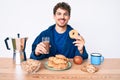 Young caucasian man with curly hair eating breakfast holding chocolate beverage and donut smiling with a happy and cool smile on Royalty Free Stock Photo