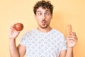 Young caucasian man with curly hair holding apple and croissant making fish face with mouth and squinting eyes, crazy and comical