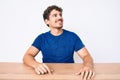 Young caucasian man with curly hair wearing casual clothes sitting on the table looking away to side with smile on face, natural Royalty Free Stock Photo