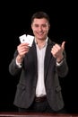 Young caucasian man wearing suit holding two aces in his hand and showing thumb up hand gesture with smile on black Royalty Free Stock Photo