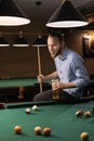 Young caucasian man drinking beer and playing billiards while sitting on the pool table contemplating hitting the ball Royalty Free Stock Photo